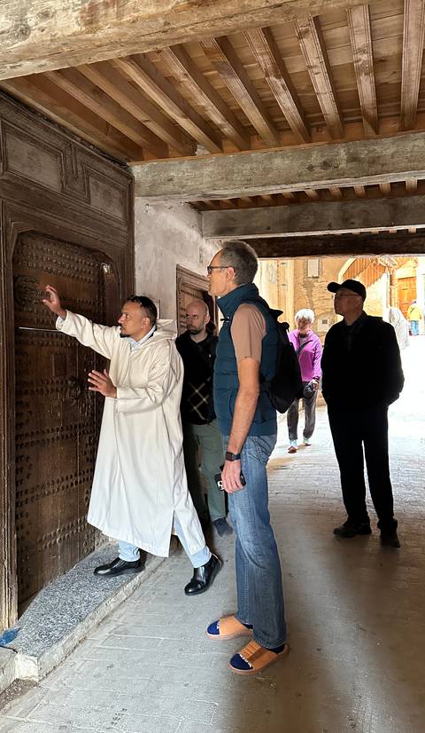       A local guide in a white djellaba shows a carved wooden door to a small group of tourists inside a narrow Moroccan medina passage.
  
