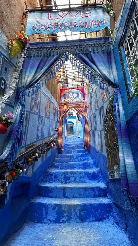       Vibrant blue-painted stairway in Chefchaouen decorated with paintings, tassels and hanging lights.
  