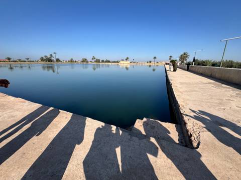       Large rectangular reflecting pool at Menara Gardens with tourist shadows cast on concrete walkway under clear blue sky.
  
