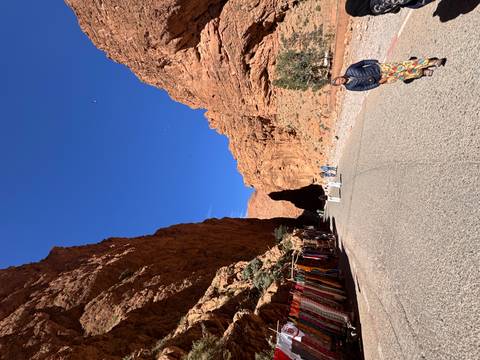       People walk along road through towering red cliffs of Todra Gorge with small market stalls on side.
  