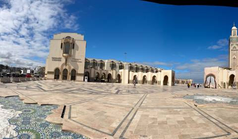       Expansive courtyard of Hassan II Mosque complex with mosaic foreground and minaret under bright blue sky.
  