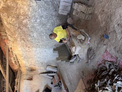       Worker chiseling clay bricks inside a rustic workshop with stacks of fresh tiles.
  