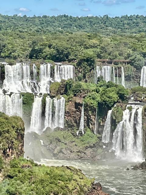       Panoramic view of multiple tiers of Iguazu Falls dropping through lush jungle.
  
