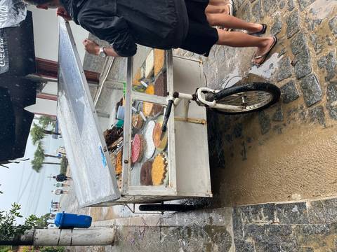       Street vendor cart displaying assorted pastries under a wet awning on a rainy cobblestone lane.
  