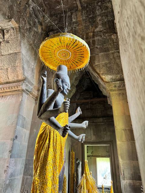       Multi-armed stone deity statue draped in a yellow cloth beneath a decorative parasol inside a temple corridor.
  
