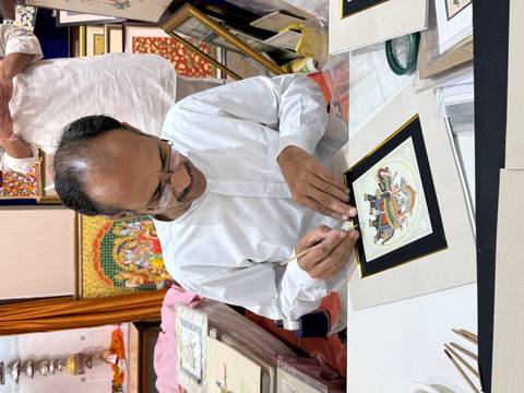       An artist in traditional attire meticulously painting a miniature artwork at a table.
  
