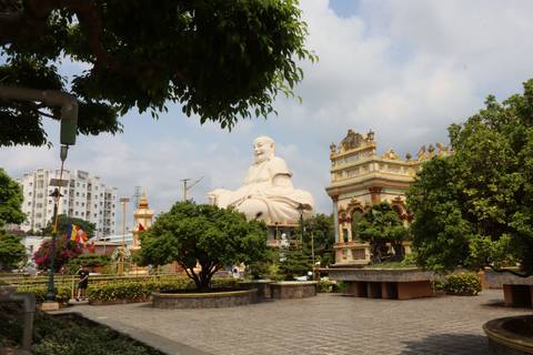       Giant smiling Buddha statue seated in a lush garden courtyard under a hazy sky.
  