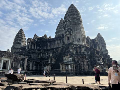       Tourists photographing the massive stone towers and steep steps of Angkor Wat under blue sky.
  
