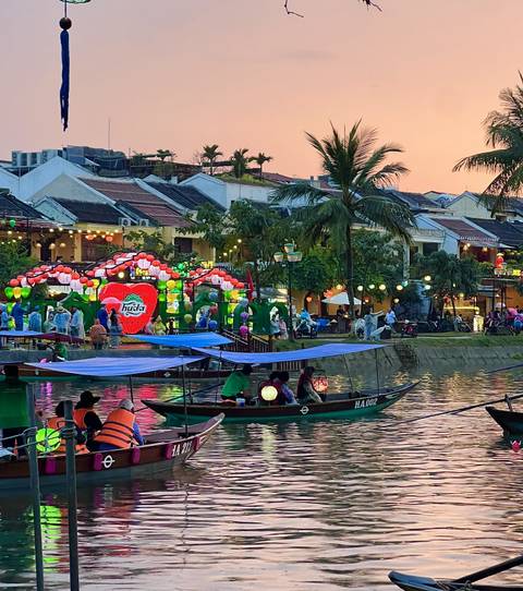       Lantern-lit boats and festive riverside market in Hoi An at dusk with colorful lights reflecting on the water.
  