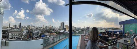       Panoramic rooftop view over Bangkok skyline with guests relaxing by an infinity pool under scattered clouds.
  