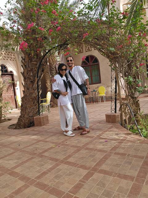       Couple in breezy white outfits embrace in shaded courtyard of Moroccan riad.
  