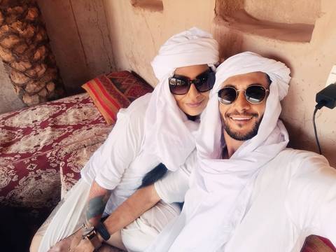       Couple relax on patterned cushions wearing white turbans and sunglasses in desert lodge.
  