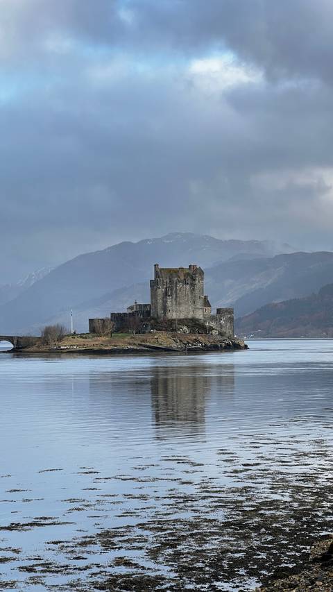       Iconic Eilean Donan Castle stands on a small island surrounded by still water against misty Highland mountains.
  
