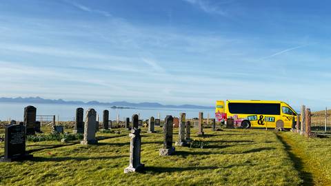       A bright yellow tour minibus parked beside an old cemetery overlooking a calm sea and distant islands.
  