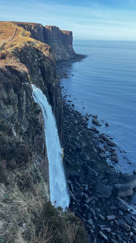       A dramatic waterfall plunges from basalt cliffs straight into the sea with scattered dark rocks below.
  