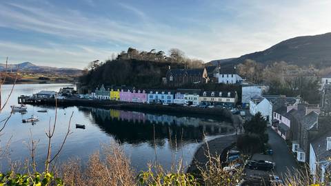       Colourful harbour cottages of Portree reflect in calm waters with surrounding hills under a clear sky.
  
