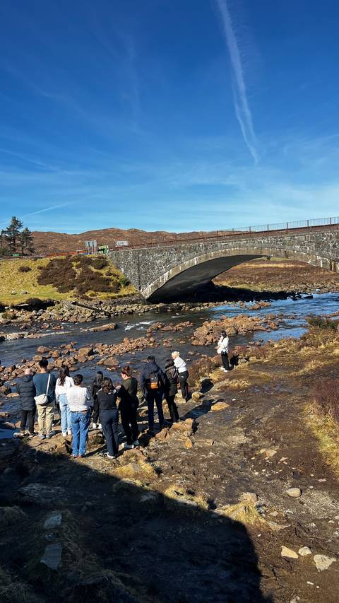       Tourists gather near an old stone arch bridge spanning a rocky Highland stream under bright sun.
  