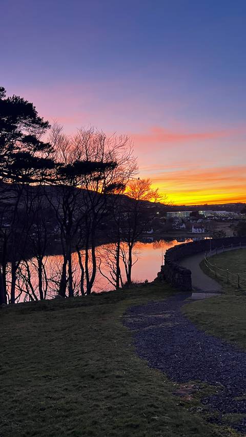       A fiery orange sunset reflects on a quiet loch framed by dark silhouetted trees and rolling hills.
  