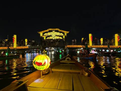       Night boat ride on lantern-lit river in Hoi An with illuminated bridge and reflections.
  