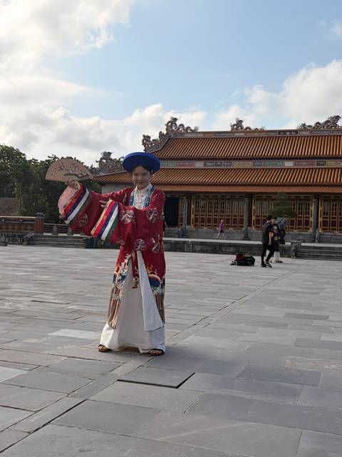       Performer in red traditional Vietnamese costume posing with a fan in an imperial courtyard
  