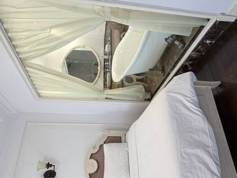       Elegant hotel room with white bed adjacent to bathroom featuring clawfoot tub behind glass wall
  