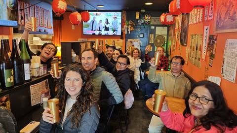       Happy travelers raise cups inside a cozy Japanese izakaya decorated with lanterns and posters.
  