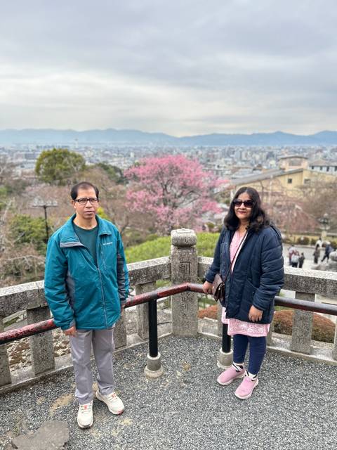       Couple posing beside a blossoming cherry tree overlooking Kyoto city from a temple terrace.
  