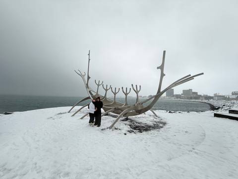       Two travellers stand on snowy ground beside the Sun Voyager sculpture with Reykjavik's harbour skyline in fog.
  