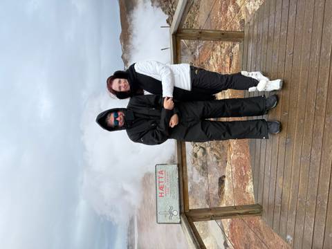       Bundled travellers pose on a wooden platform amid billowing geothermal steam and warning signs.
  