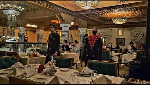       Dimly lit Moroccan restaurant with ornate décor, musicians and diners at candle-lit tables.
  