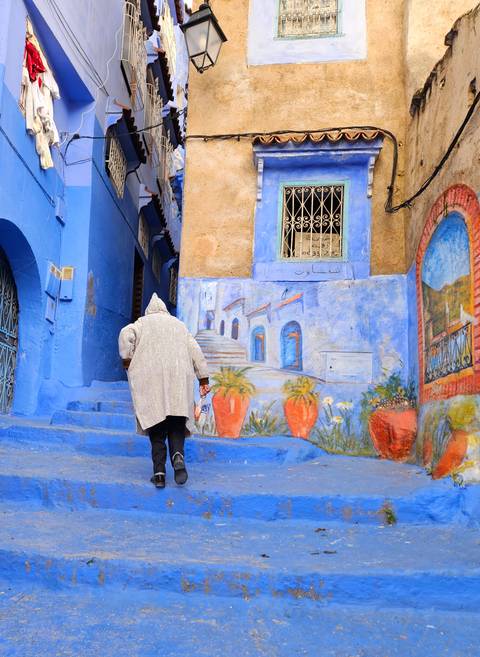       Elderly person in hooded cloak ascending blue steps beside colorful mural in Chefchaouen.
  