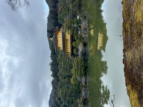       Golden Pavilion temple reflecting in a calm pond surrounded by pine trees under overcast skies.
  