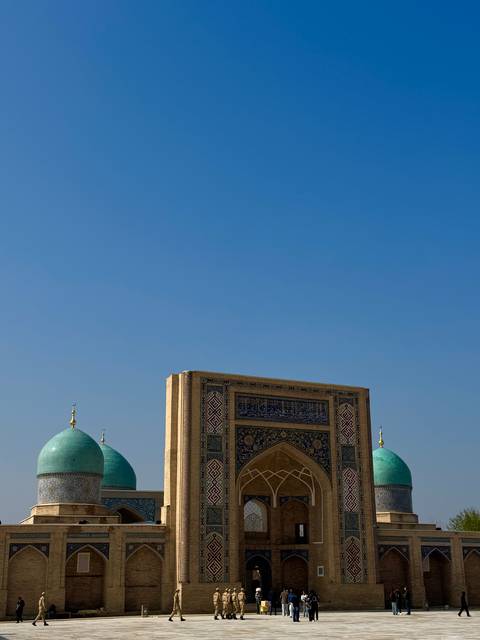       Blue domes and an ornate portal of a historic Islamic complex stand beneath a vast clear sky.
  