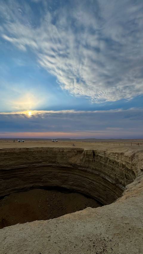       Cars and tiny figures stand at the rim of a massive desert sinkhole under a dramatic sky.
  