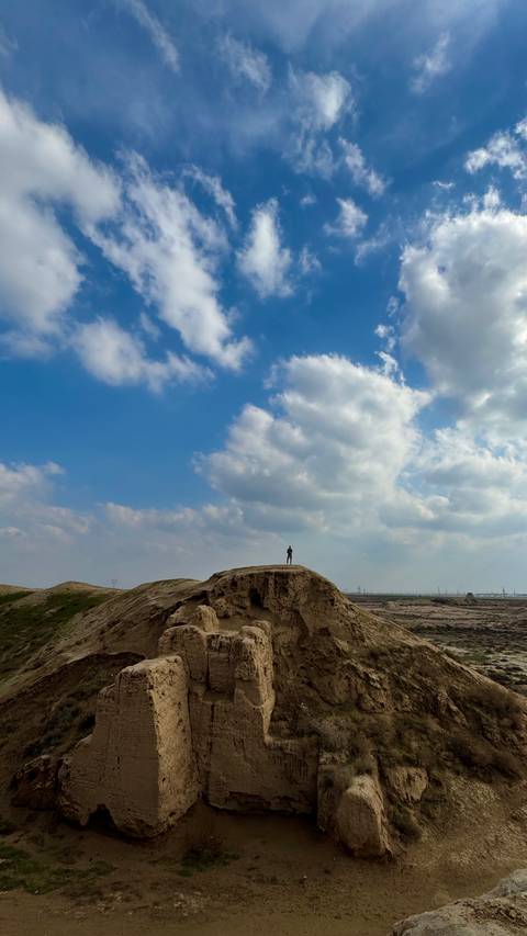       A lone traveler stands atop a sandstone hill beneath a vibrant blue sky filled with billowing clouds.
  