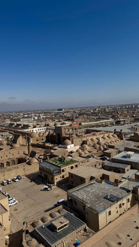       An elevated panoramic view of a historic mud-brick city with domes, minarets, and surrounding modern sprawl.
  