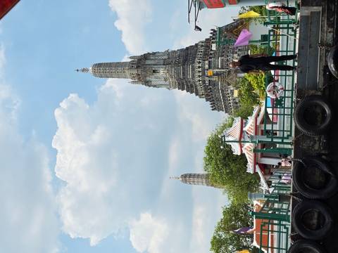       Iconic prang of Wat Arun rises over a riverside pier under a blue sky with fluffy clouds and visitors on the dock
  
