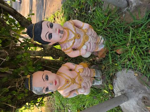       Two painted clay child statues in traditional dress standing on grass beside a small tree
  