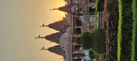       Cluster of ornate temple spires silhouetted by a golden sunset behind manicured gardens
  