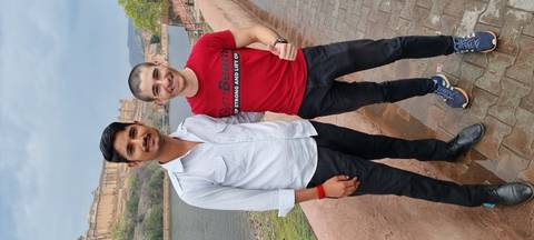       Two travelers smiling for a photo on a riverside walkway with a historic fort rising on the hill behind
  