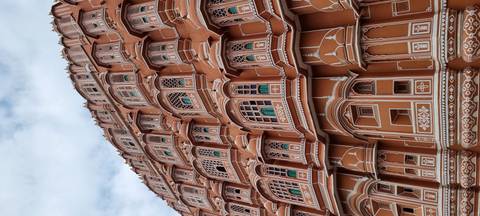       Detailed façade of the pink sandstone Hawa Mahal with rows of ornate windows against a cloudy sky
  