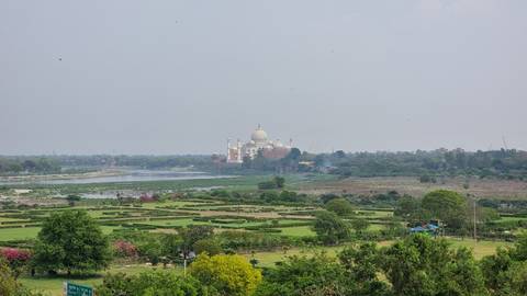       Distant view of the Taj Mahal across a broad river plain surrounded by green gardens and trees under hazy sky
  