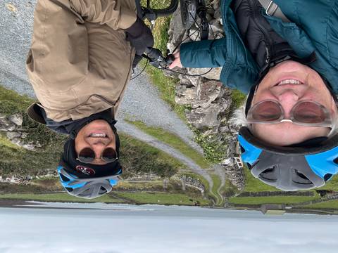       Two cyclists wearing helmets smile for a selfie on a narrow country lane bordered by stone walls with green fields and the sea in the background
  