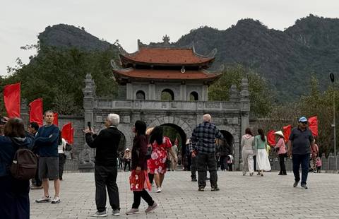       Tourists holding red flags gather before an ancient stone gate with a red-tiled roof set against karst mountains in Ninh Binh province
  