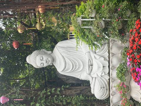       White seated Buddha statue in a lush garden decorated with lanterns and colorful flowers
  