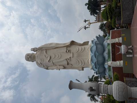       Giant standing Buddha atop a lotus pedestal reaches into a partly cloudy sky beside temple grounds
  