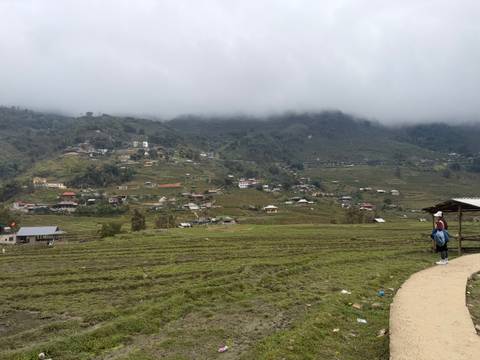       Mist-covered valley with scattered houses and terraced fields viewed from a pathway where a traveller pauses with a backpack
  