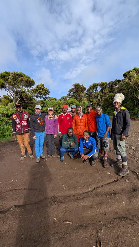       Hikers and porters pose together on a forested trailhead with lush greenery in the background.
  