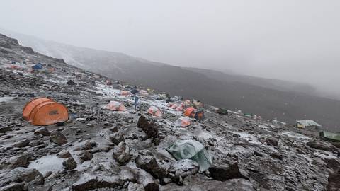       A high-altitude campsite with dozens of orange tents scattered over a rocky, lightly snow-dusted slope in misty weather.
  
