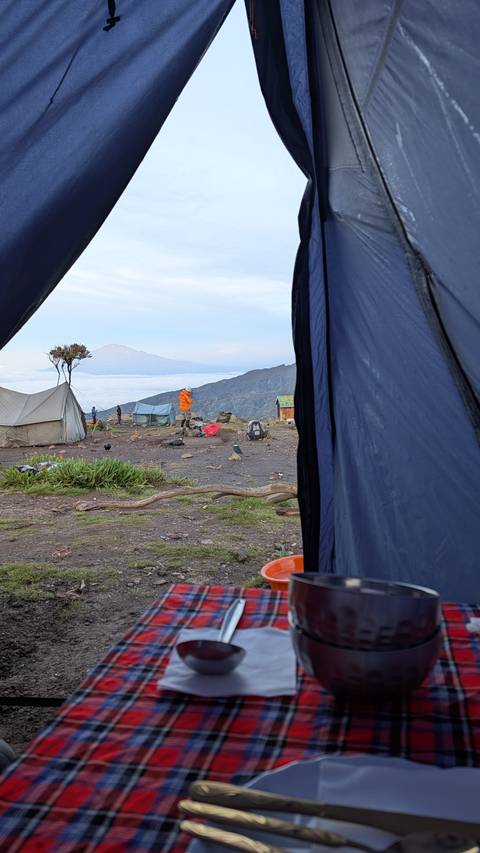       View from inside a tent onto a high mountain campsite above a sea of clouds with a distant volcanic summit.
  